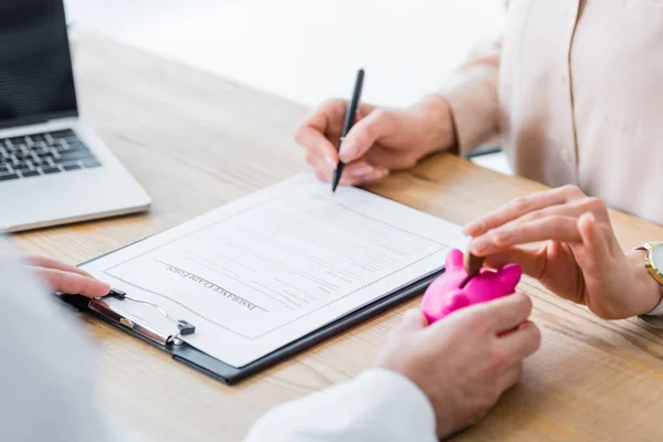 Partial view of doctor holding piggy bank in hand while woman holding coin and signing document — Stock Photo