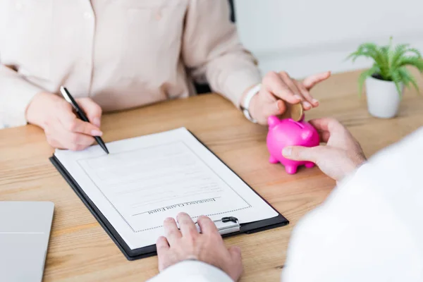 Cropped view of doctor holding piggy bank in hand while woman holding coin and signing document — Stock Photo