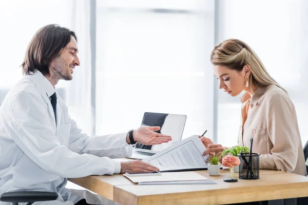 Mujer firmar formulario de reclamación de seguro durante la cita con el médico en la clínica - foto de stock