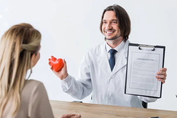 Selective focus of happy doctor holding red heart and insurance claim form in hands while looking at patient — Stock Photo