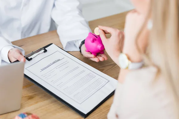 Vue recadrée du médecin tenant le formulaire de demande de règlement d'assurance et tirelire à la main tandis que la femme tenant pièce — Photo de stock