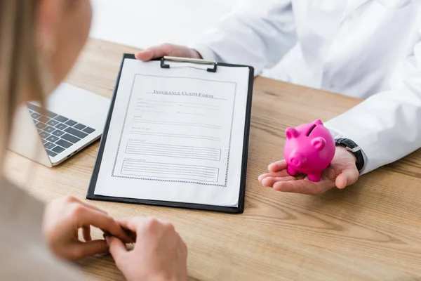 Vue recadrée du médecin tenant le formulaire de réclamation d'assurance et tirelire en main — Photo de stock