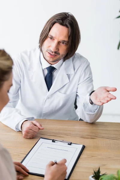Doctor talking with patient while woman signing document and sitting behind wooden table — Stock Photo