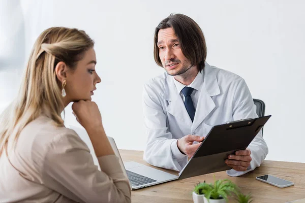 Guapo doctor sosteniendo documento en las manos, mirando paciente y sentado detrás de mesa de madera - foto de stock