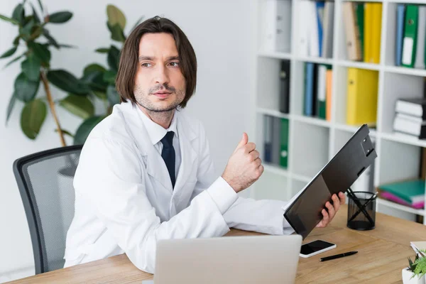 Guapo médico en brillante oficina sentado detrás de una mesa de madera, mirando hacia otro lado y sosteniendo el portapapeles en la mano — Stock Photo