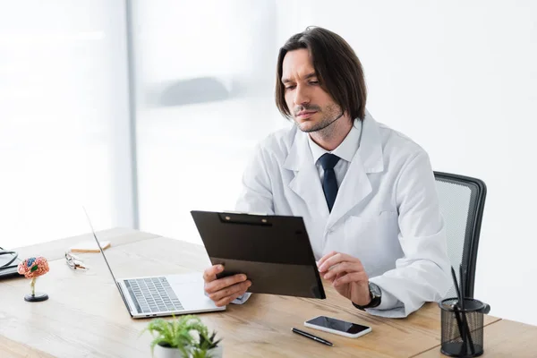 Schöner Arzt mit Klemmbrett in der Hand sitzt im Büro am Schreibtisch — Stockfoto