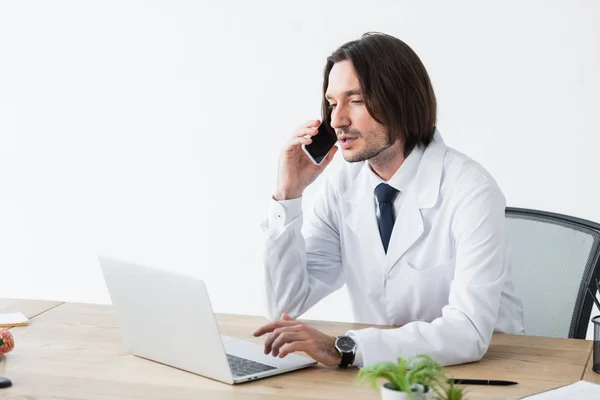 Doctor tranquilo hablando en el teléfono inteligente mientras está sentado en la oficina brillante con el ordenador portátil en la mesa - foto de stock