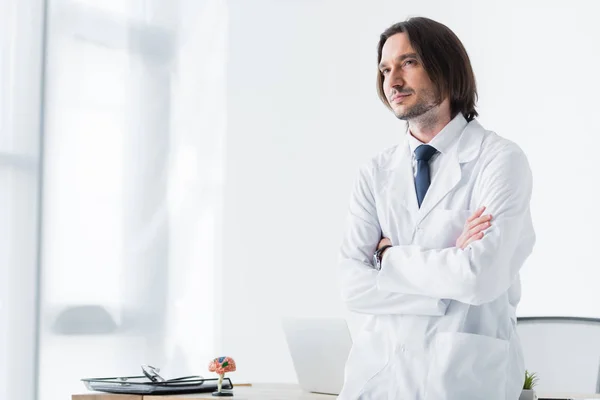 Спокойный доктор в белом халате стоит в кабинете со скрещенными руками на груди и смотрит в камеру — стоковое фото