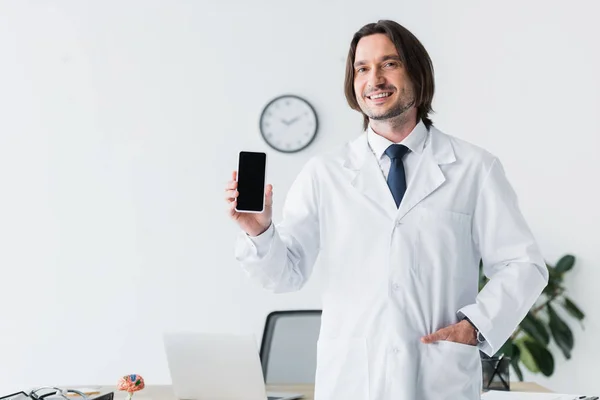 Médecin joyeux en manteau blanc regardant la caméra et tenant smartphone dans la main — Photo de stock