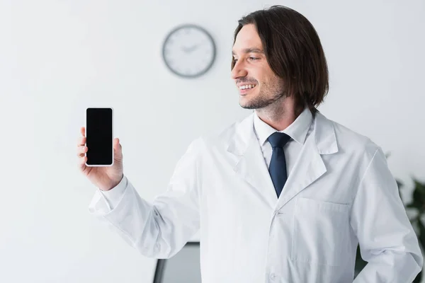 Happy doctor in white coat holding smartphone in hand and looking at display with copy space — Stock Photo