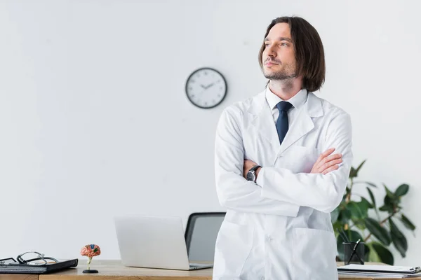 Doctor tranquilo en bata blanca con los brazos cruzados mirando hacia otro lado - foto de stock