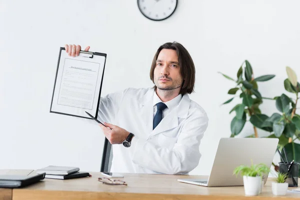 Doctor in white coat sitting behind wooden table, looking at camera and pointing with pen at medical document — Stock Photo