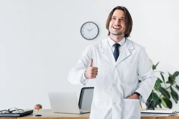 Веселий лікар у білому пальто дивиться в сторону і показує жест великого пальця вгору — стокове фото