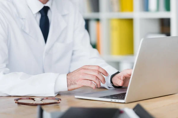 Обрезанный вид врача в белом халате, работающего с ноутбуком — стоковое фото