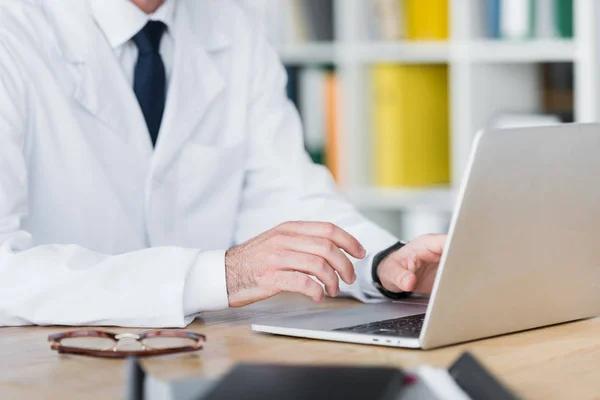 Vista recortada del médico en bata blanca trabajando con el ordenador portátil - foto de stock