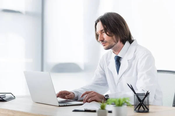 Beau médecin en manteau blanc assis derrière une table en bois et travaillant avec un ordinateur portable — Photo de stock