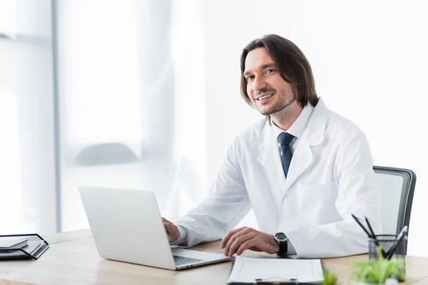 Happy doctor in white coat looking at camera while working with laptop — Stock Photo