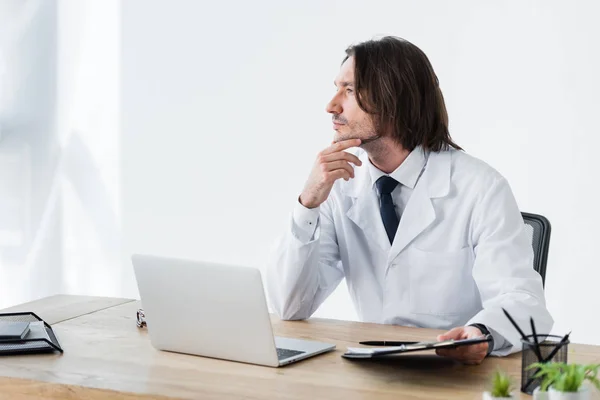 Médico de bata blanca mirando hacia otro lado mientras está sentado detrás de una mesa de madera con portátil — Stock Photo