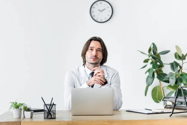 Doctor in white coat looking at camera while sitting behind wooden table with laptop — Stock Photo