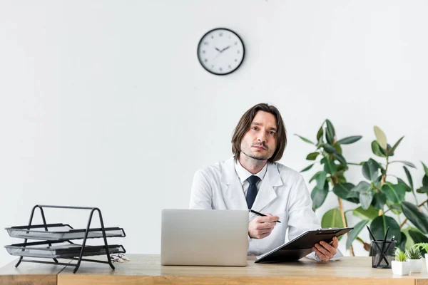 Médecin en manteau blanc regardant la caméra, tenant un stylo avec un document médical dans les mains et assis derrière une table en bois avec un ordinateur portable — Photo de stock