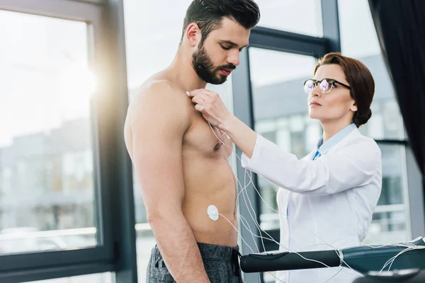 Hermoso médico poniendo electrodos en deportista sin camisa durante la prueba de resistencia - foto de stock