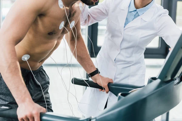 Обрезанный вид врача, надевающего электроды на спортсмена без рубашки во время теста на выносливость — стоковое фото
