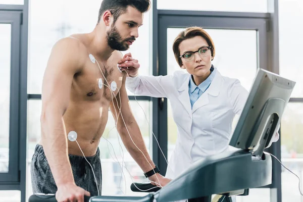 Красивый врач надевает электроды на спортсмена без рубашки во время теста на выносливость — стоковое фото