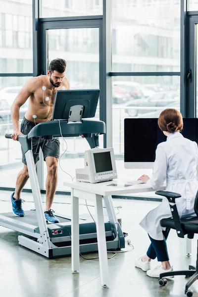Лікар сидить за комп'ютерним столом і проводить тест на витривалість зі спортсменом у спортзалі — стокове фото