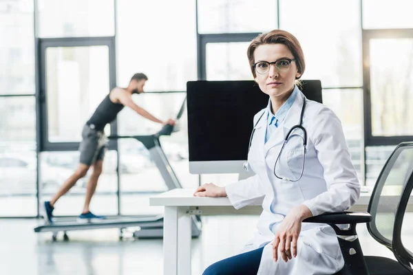 Médico sentado en el escritorio cerca de deportista corriendo en la cinta de correr durante la prueba de resistencia en el gimnasio - foto de stock