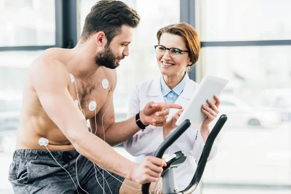 Lächelnder Arzt mit digitalem Tablet neben hemdslosem Sportler beim Härtetest — Stockfoto