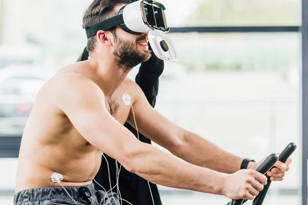 Smiling shirtless desportista em realidade virtual headset com eletrodos trabalhando em elíptico durante teste de resistência no ginásio — Fotografia de Stock