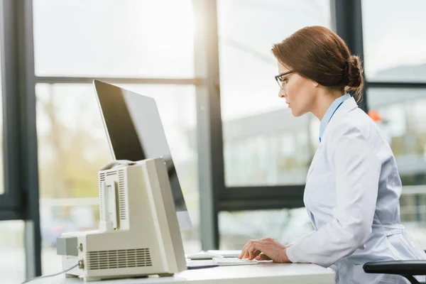 Schöne Ärztin im weißen Mantel sitzt am Computer-Schreibtisch — Stockfoto