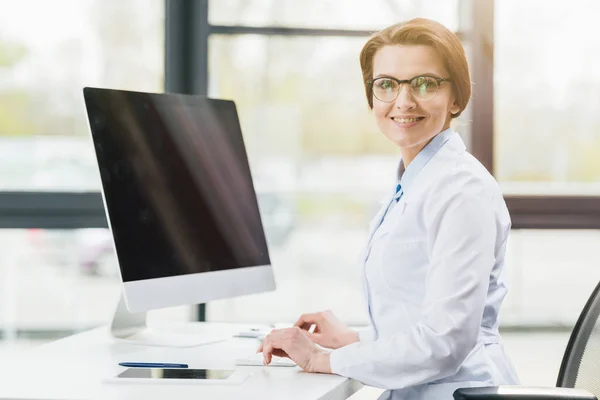 Beautiful happy doctor in white coat sitting near computer with blank screen — Stock Photo