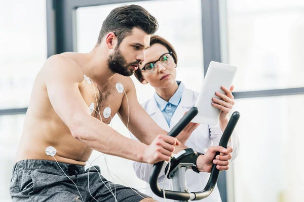 Doctor with digital tablet near shirtless sportsman during endurance test in gym — Stock Photo