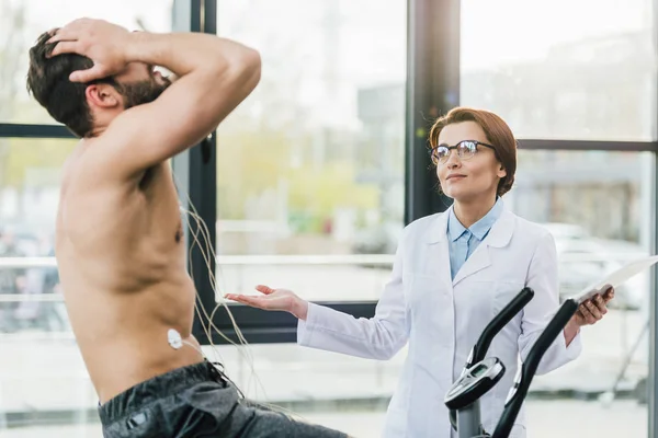 Médecin avec tablette numérique gestuelle près sportif torse nu lors d'un test d'endurance dans la salle de gym — Photo de stock