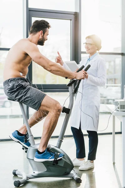 Sportsman training on elliptical machine near doctor with digital tablet during endurance test — Stock Photo