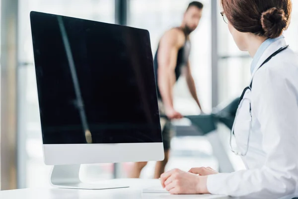 Selective focus of doctor sitting near computer with blank screen in gym — Stock Photo