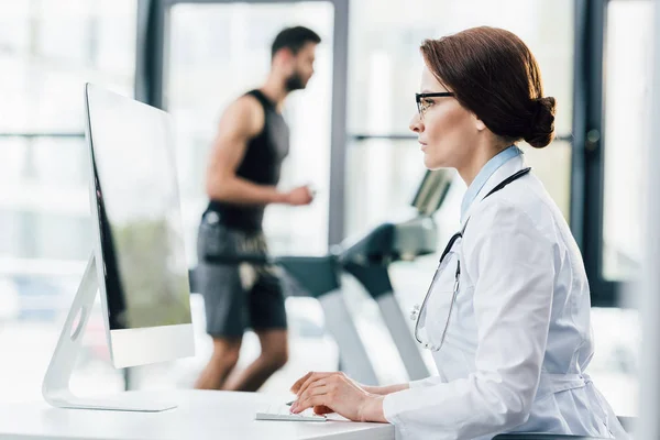 Doctor en gafas usando computadora mientras deportista corriendo en la cinta de correr durante la prueba de resistencia en el gimnasio - foto de stock