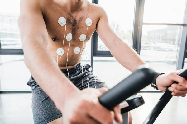 Cropped view of sportsman with electrodes training on elliptical during endurance test in sports center — Stock Photo