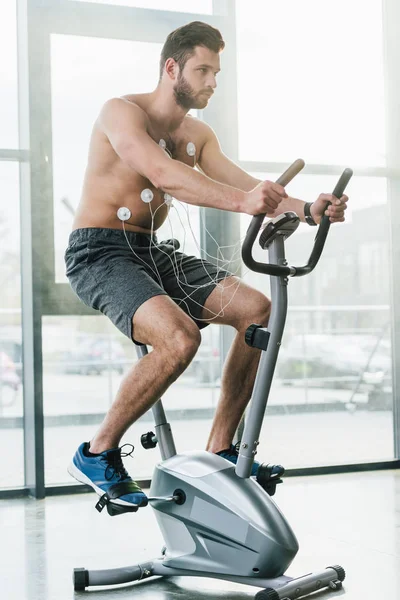 Sportif avec électrodes d'entraînement sur elliptique lors d'un test d'endurance en salle de gym avec lumière du soleil — Photo de stock