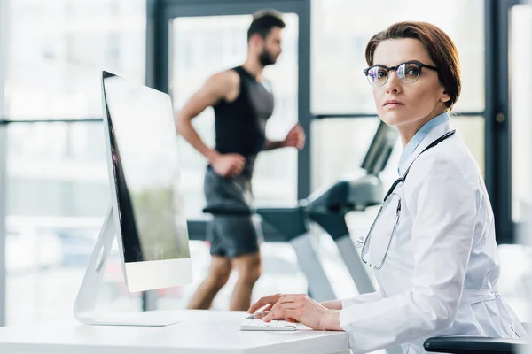 Doctor in glasses using computer while sportsman running on treadmill during endurance test in gym — Stock Photo