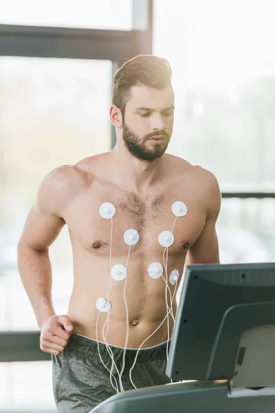 Handsome sportsman with electrodes running on treadmill during endurance test in gym with daylight — Stock Photo