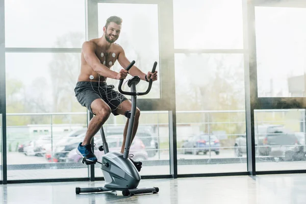 Smiling handsome sportsman with electrodes on elliptical during endurance test in gym — Stock Photo