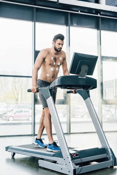 Apuesto deportista muscular con electrodos corriendo en la cinta de correr durante la prueba de resistencia en el gimnasio - foto de stock