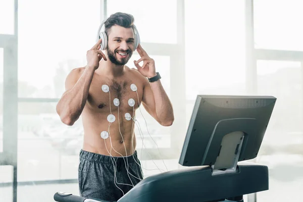 Sonriente deportista muscular en auriculares corriendo en cinta de correr durante la prueba de resistencia en el gimnasio - foto de stock