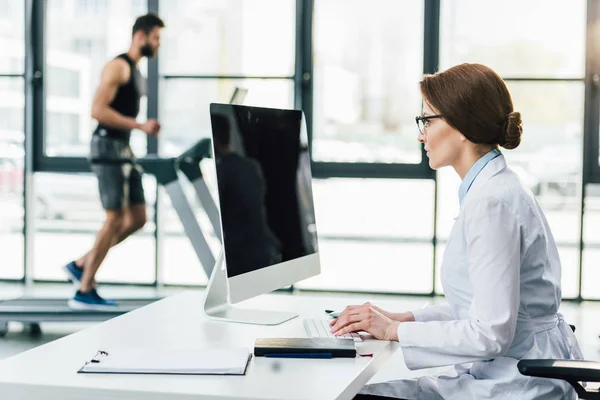 Médico usando computador enquanto desportista correndo em esteira rolante durante teste de resistência no ginásio — Stock Photo