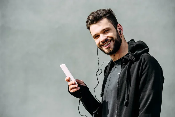 Hombre sonriente con capucha y auriculares usando teléfono inteligente en gris y mirando a la cámara - foto de stock