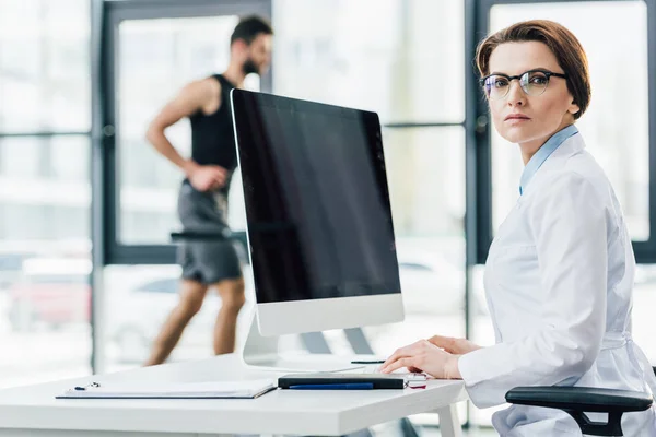 Selective focus of beautiful doctor sitting near computer with blank screen in gym — Stock Photo