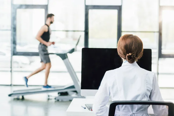 Médico que utiliza la computadora mientras que el deportista que corre en la cinta de correr durante la prueba de resistencia en el gimnasio - foto de stock