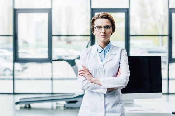 Doctor in white coat with crossed arms looking at camera at gym — Stock Photo